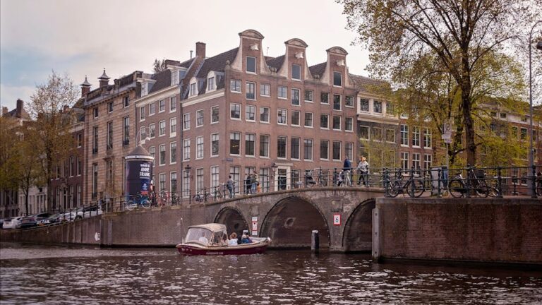 Gracht in Amsterdam met bakstenen brug, rij van fietsen, historische gevelhuizen en bootje in het water.